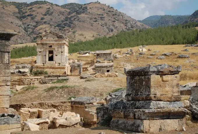 A pedimented temple tomb stands among fallen blocks on the golden grass of the Hierapolis necropolis below wooded hills.