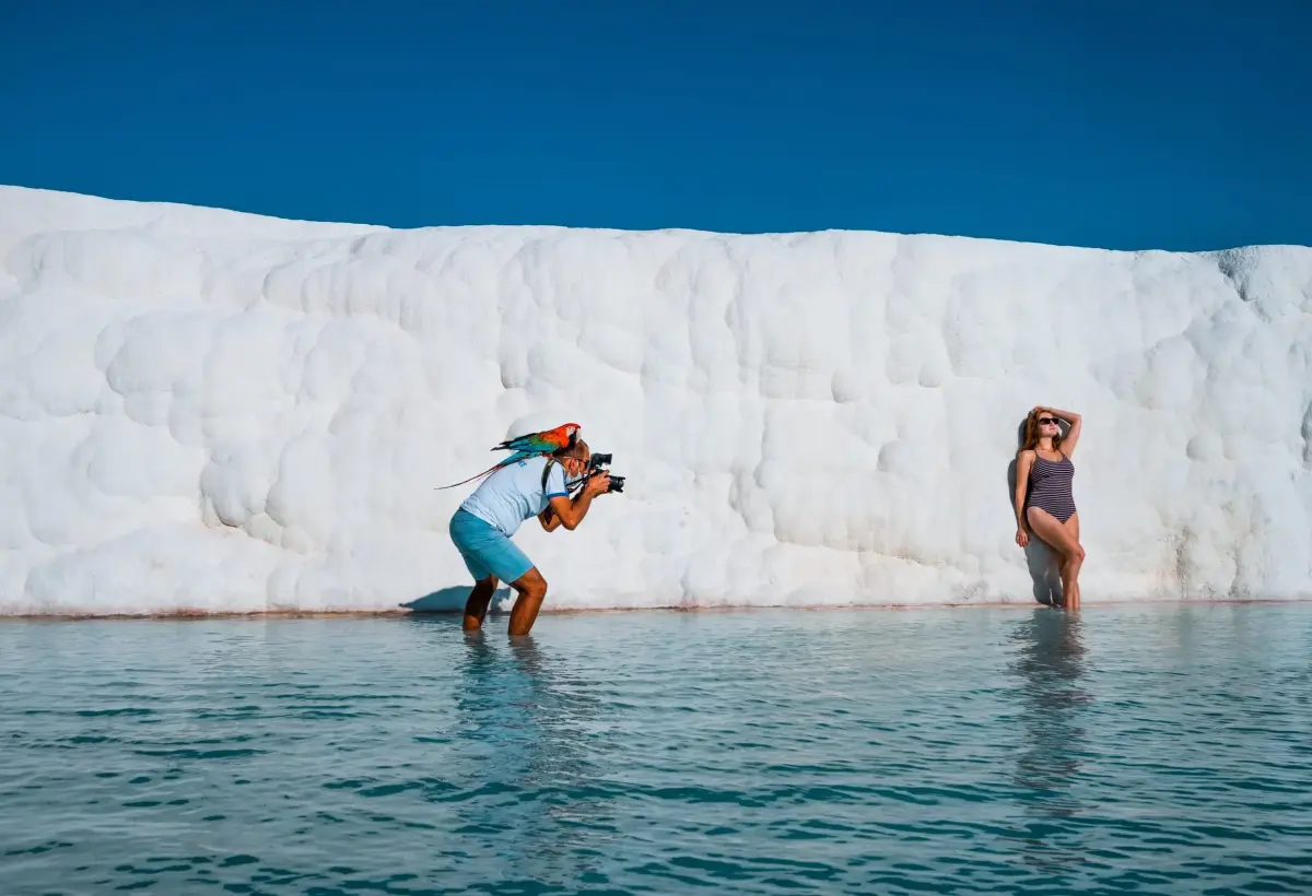 A photographer wades in shallow water at Pamukkale to shoot a woman posing against the towering white travertine wall.