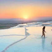 A bather walks through ankle-deep water on the Pamukkale terraces as the sun sets over the wide plain ahead.