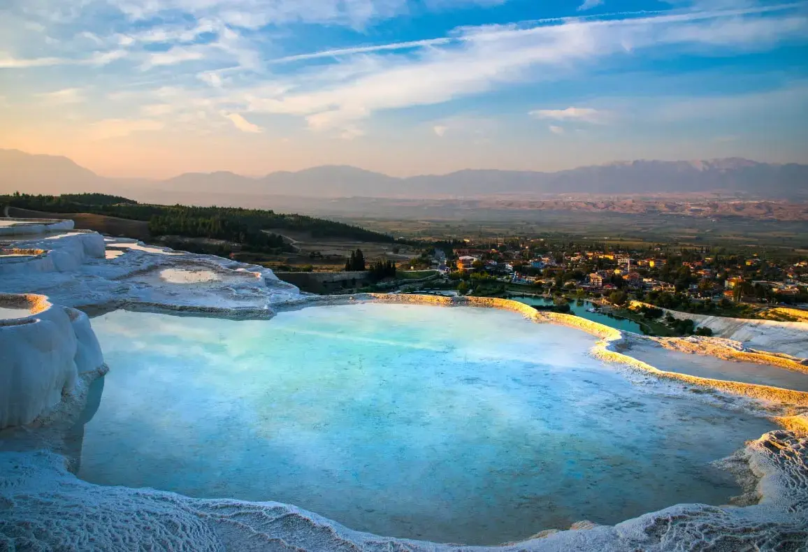 A broad pale blue pool fills the travertine at Pamukkale with the town and the distant mountains lit at dusk.