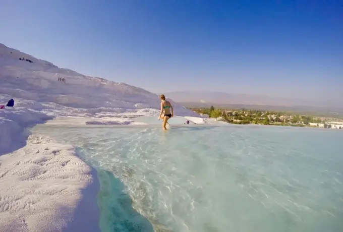 A bather stands at the edge of a wide shallow pool at Pamukkale with the white hillside and green valley beyond.