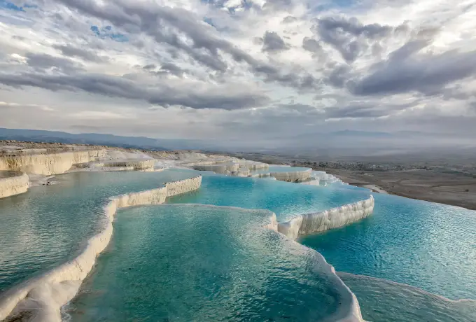 Turquoise pools sit in the white travertine at Pamukkale under a low bank of heavy grey cloud.