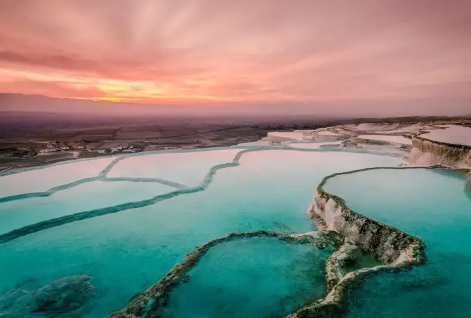Teal thermal pools fill the terraced basins at Pamukkale beneath a soft pink and orange sunset sky.