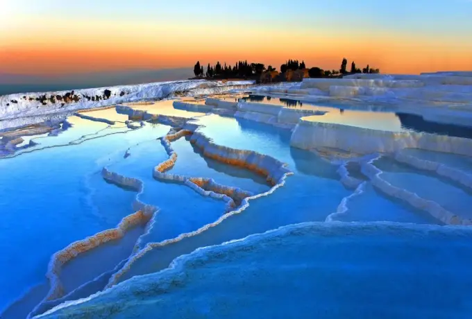 Deep cobalt water fills the terrace pools at Pamukkale at dusk with cypress trees standing on the ridge above.