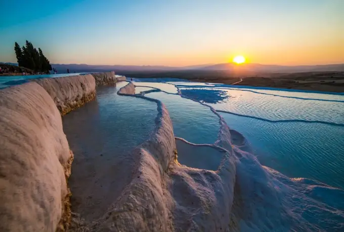 A long shallow channel of water runs between white travertine ledges at Pamukkale with the sun low over the valley.
