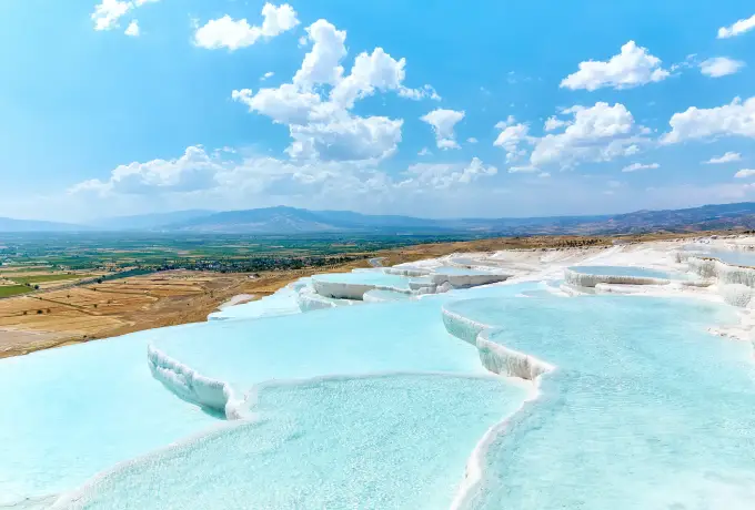 White travertine ledges and pale blue pools spread across the Pamukkale hillside under a blue sky with white clouds.