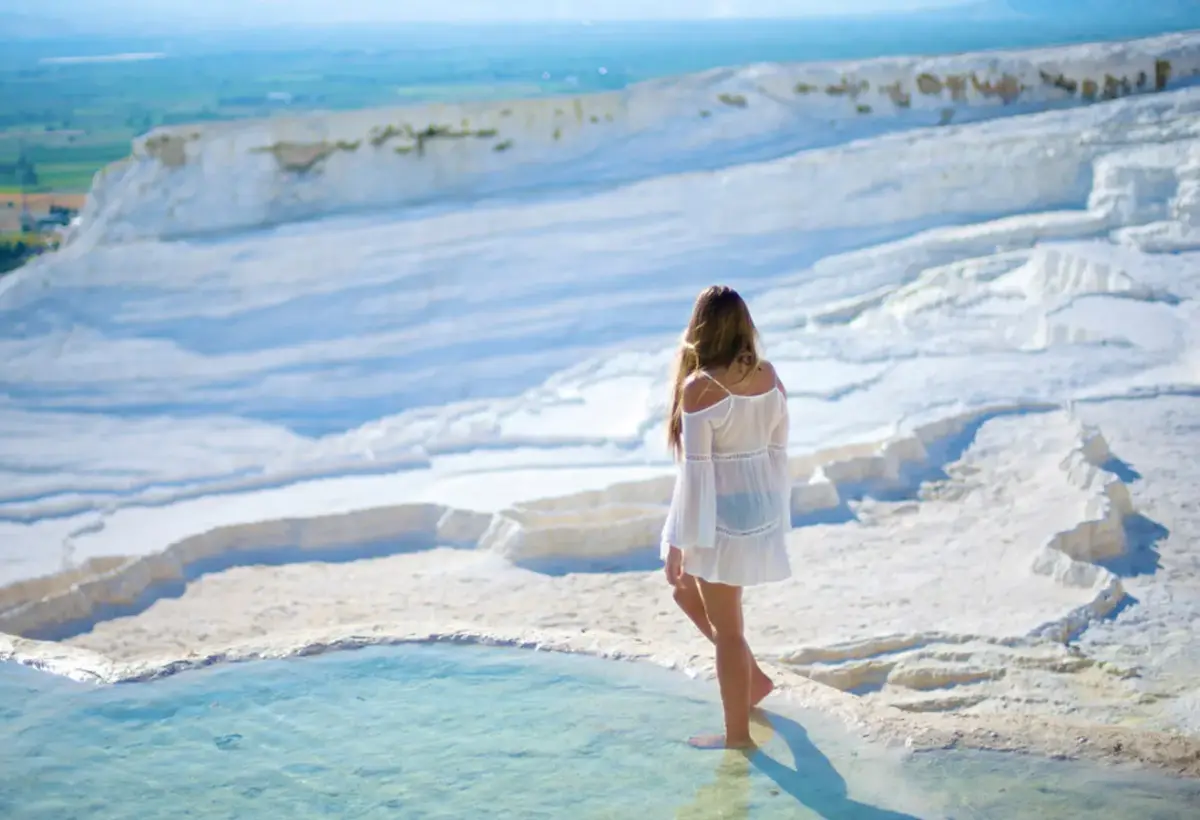 A woman in a white dress stands barefoot on the edge of a travertine pool at Pamukkale above the wide green valley.
