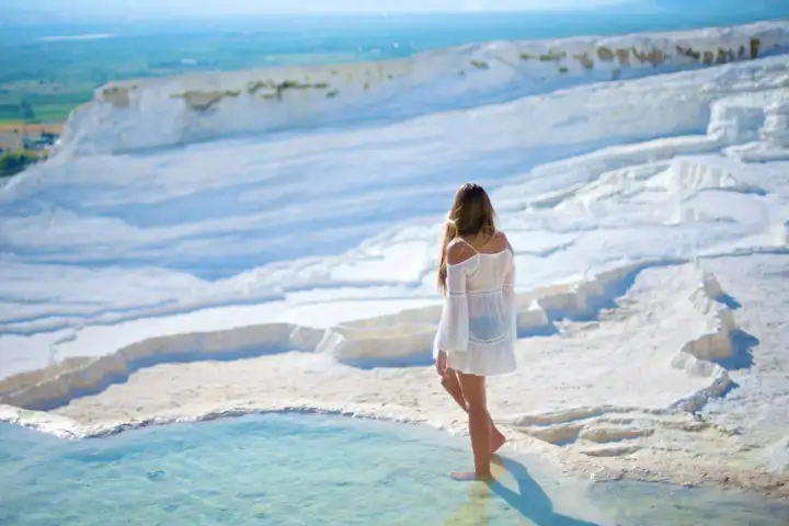 A woman in a white dress stands barefoot on the edge of a travertine pool at Pamukkale above the wide green valley.