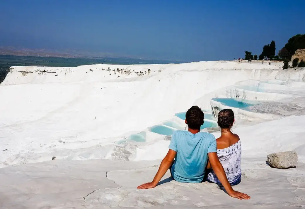 A couple sits together on the white travertine at Pamukkale looking out over the stepped pools and the valley.