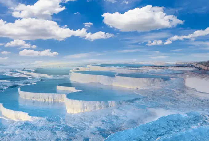 A long white travertine ledge drops into a broad pale blue pool at Pamukkale under a sky of round white clouds.