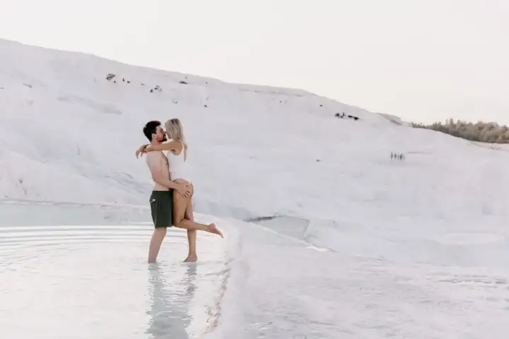 A couple embraces in ankle-deep water at Pamukkale with the vast smooth white travertine slope rising behind them.