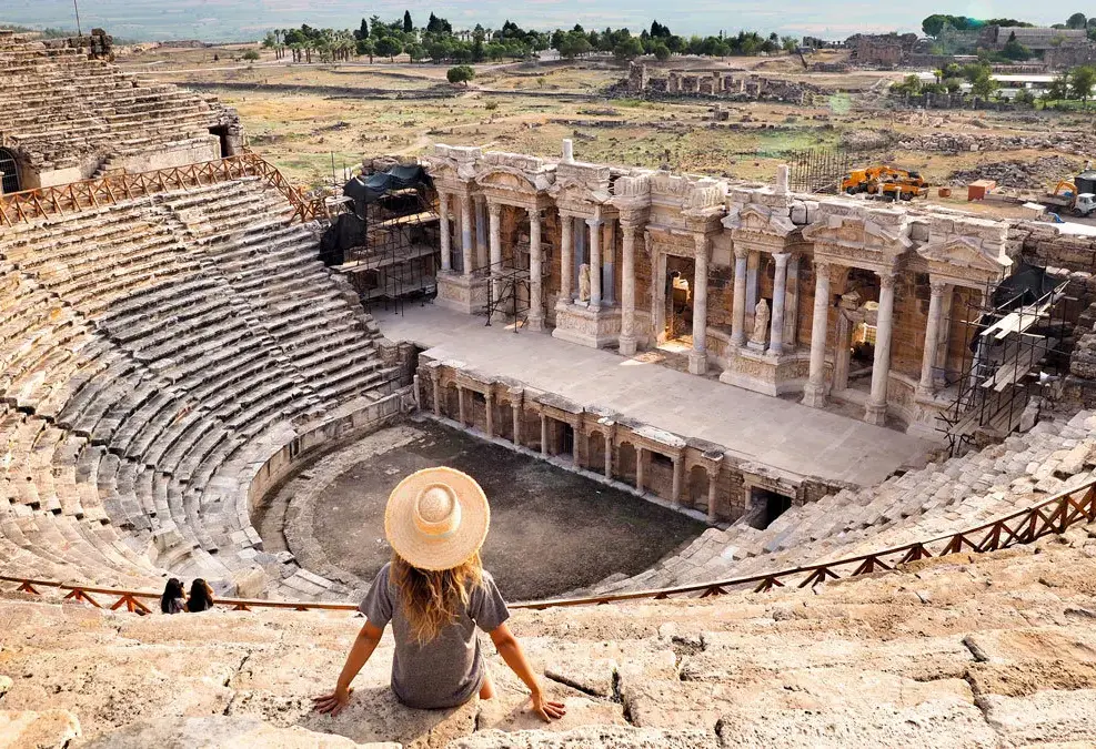 A visitor in a wide sun hat sits on the stone tiers of the Hierapolis theatre looking down at the columned stage.