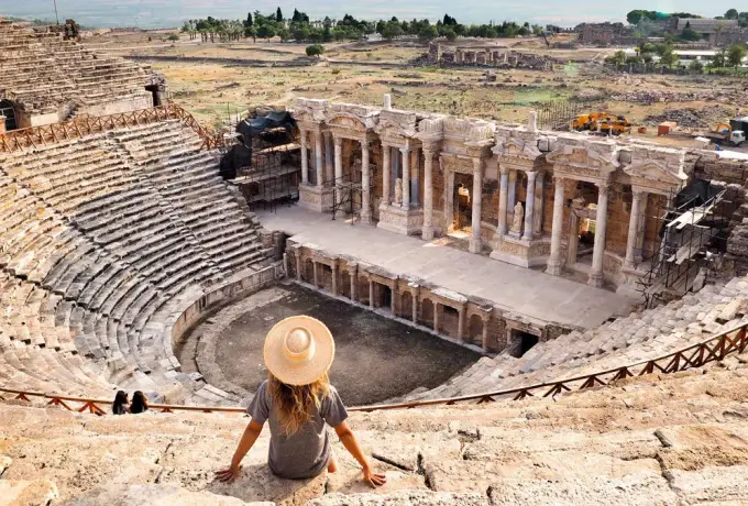 A visitor in a wide sun hat sits on the stone tiers of the Hierapolis theatre looking down at the columned stage.