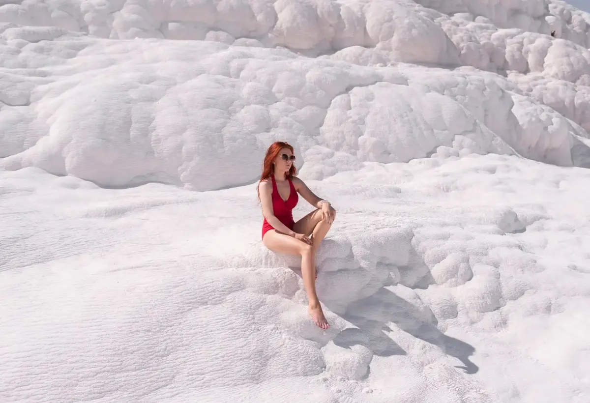 A visitor in a red swimsuit sits on the rippled white travertine at Pamukkale in bright midday light.