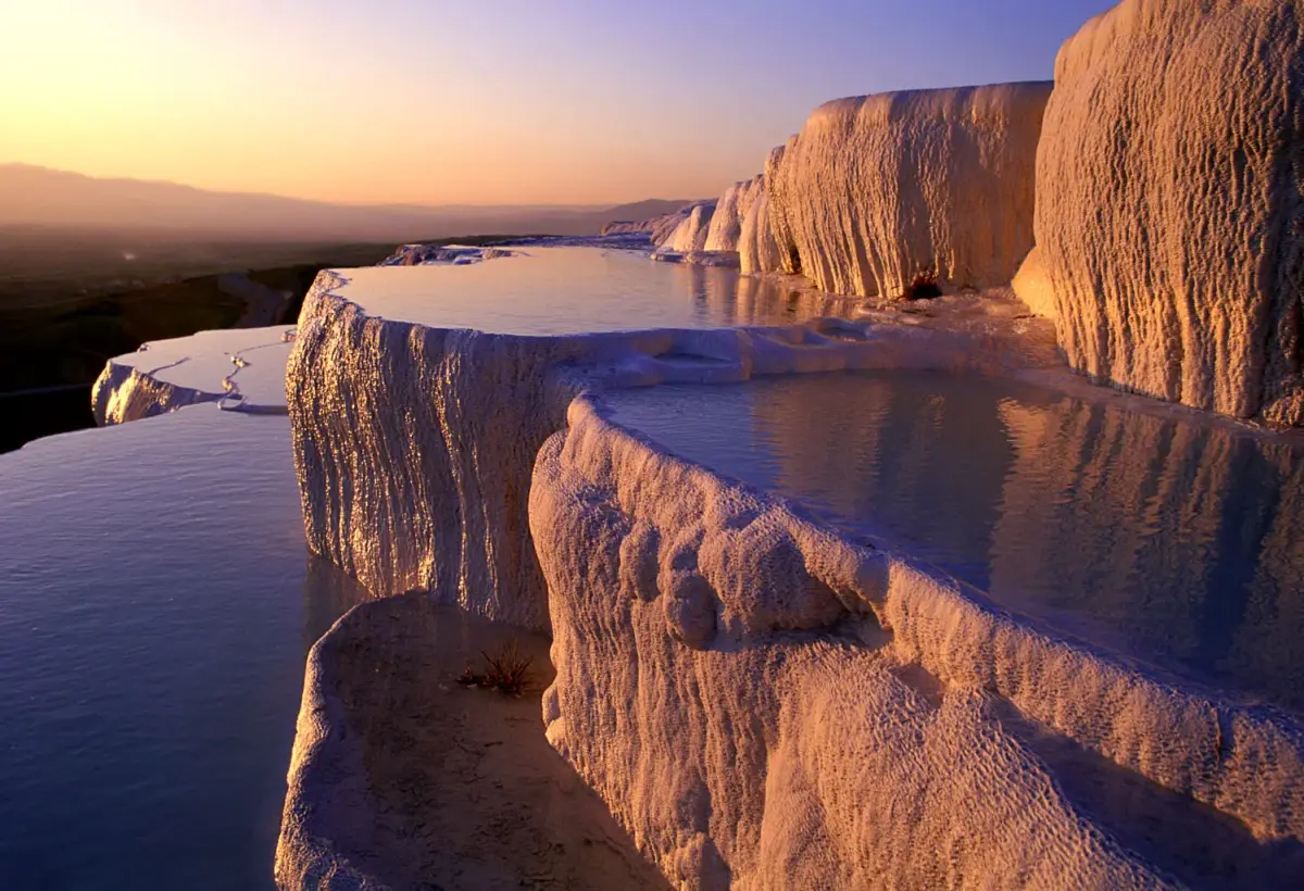 Chalky travertine cliffs catch warm light at dusk at Pamukkale while mist drifts off the pools below.