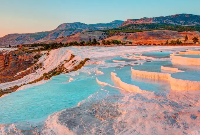 Scalloped white basins hold pale turquoise water at Pamukkale as soft sunset light warms the hills behind.