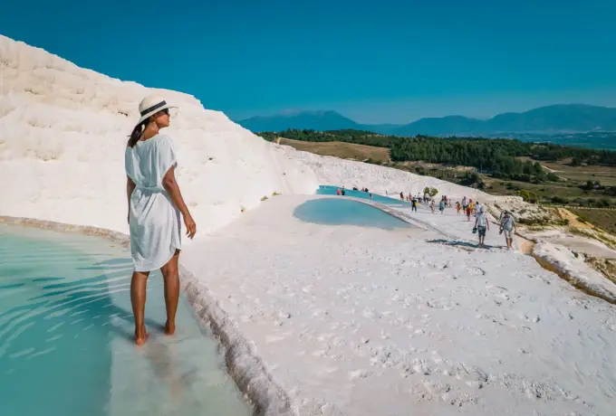 A woman in a white dress and sun hat walks the wet travertine at Pamukkale with visitors along the ridge behind.