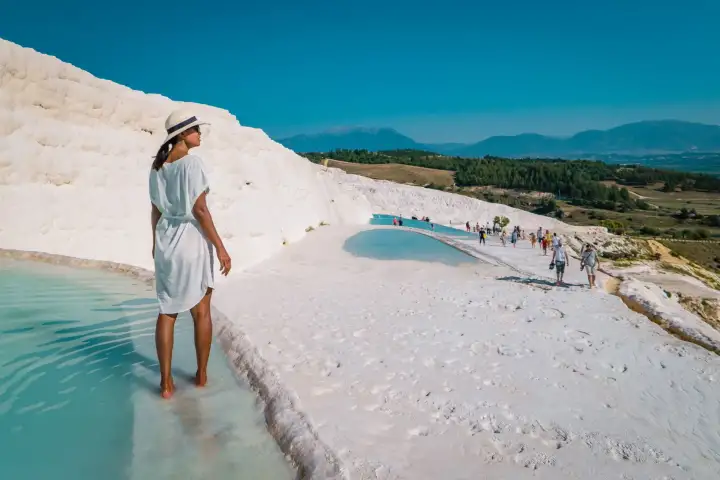 A woman in a white dress and sun hat walks the wet travertine at Pamukkale with visitors along the ridge behind.