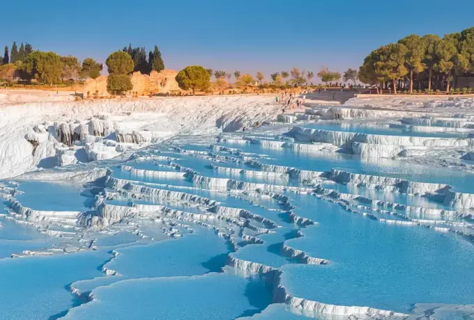 Rows of narrow white ledges hold blue water on the Pamukkale hillside with pines and ruins along the upper plateau.