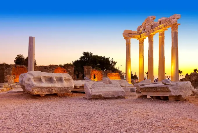 The Temple of Apollo at Side glows in warm dusk light above overturned sarcophagi and carved stone blocks spread across the seafront archaeological site.