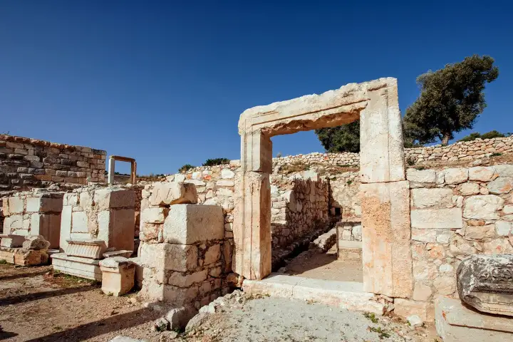 A tall marble doorway with a heavy lintel stands among the low rubble walls of ancient Troy under a deep blue sky.