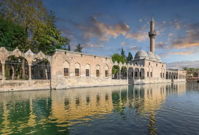 The mosque and minaret at Balikligol are mirrored in the still pool at golden hour, the long stone colonnade lit warm along the bank.