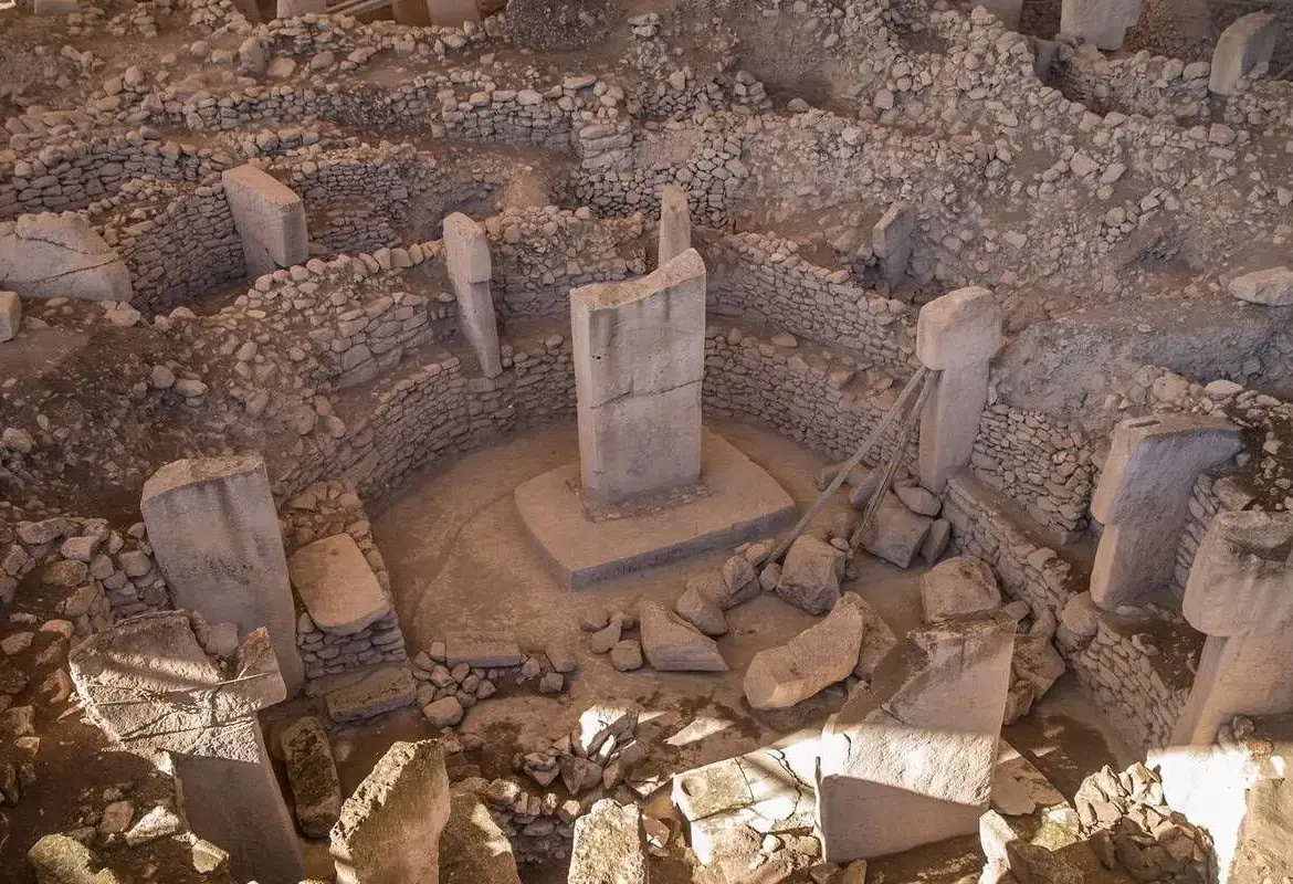 An overhead view of a Gobeklitepe enclosure shows two central T-shaped pillars on a raised base inside a ring of shorter stones.