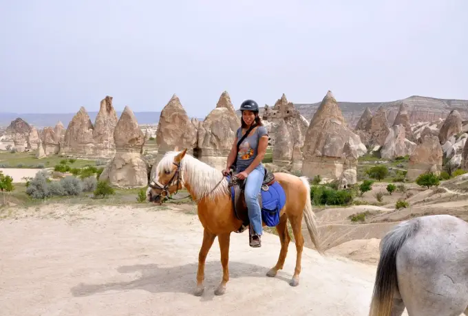 A rider sits on a palomino horse on a sandy track in Cappadocia with a field of pale rock cones spread out behind.