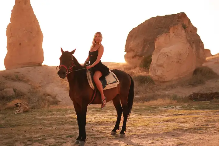 A woman sits on a brown horse in warm low sunlight between two weathered rock cones in a Cappadocia valley.