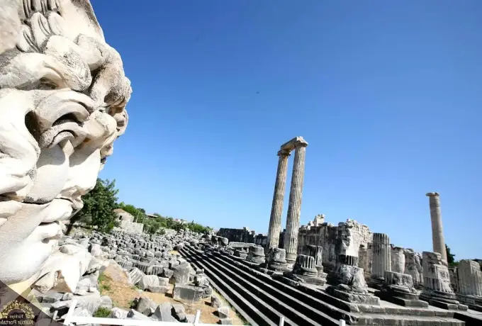 A carved Medusa head lies in the foreground beside the long stepped platform and standing columns of the sanctuary at Didyma.