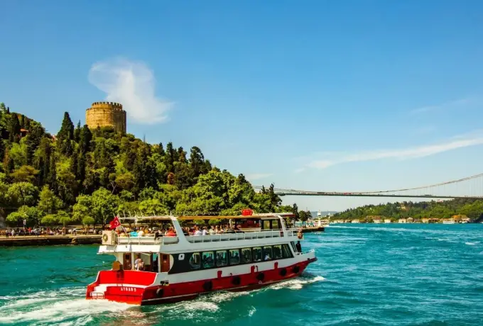 A red passenger ferry crosses the turquoise Bosphorus in Istanbul below the wooded slope and towers of the old fortress.