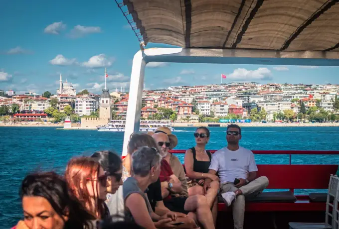 Guests sit on red benches under the canopy of a Bosphorus cruise boat in Istanbul as the shoreline slides past.