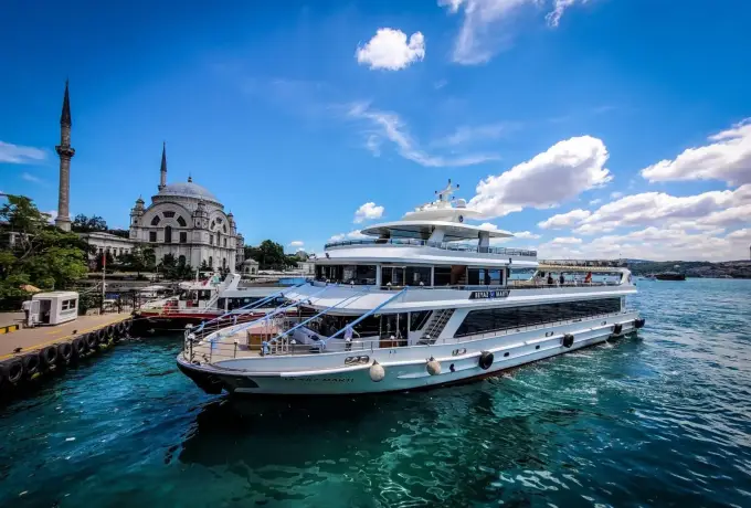 A large white cruise yacht lies at its quay on the Bosphorus in Istanbul beside a stone mosque and its minaret.