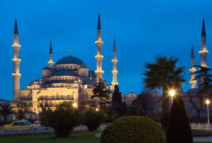 The Blue Mosque in Istanbul glows under floodlights against a deep blue night sky, seen across the dark park below.