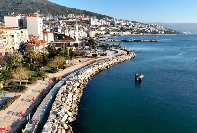 A seafront promenade at Gemlik near Bursa curves along a rock breakwater, apartment blocks lining the shore and a small boat on the calm blue water.
