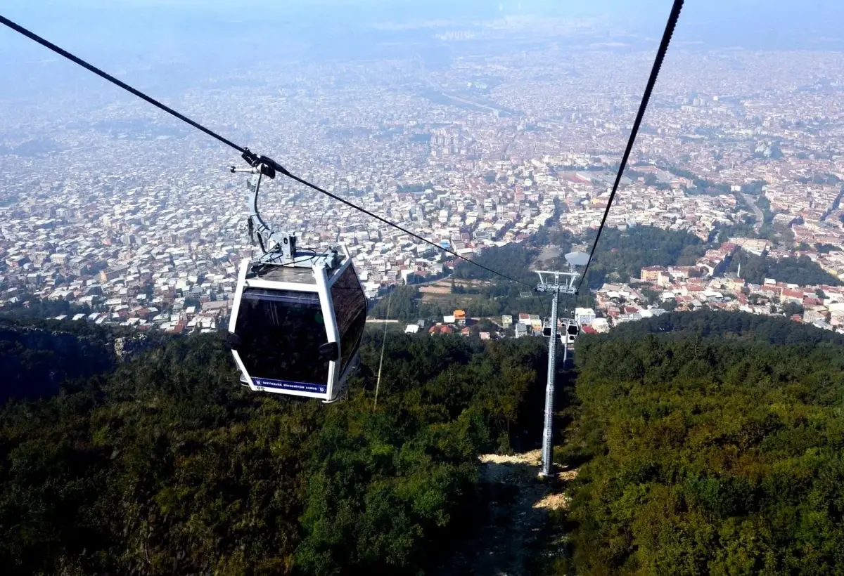 A gondola of the Bursa teleferik hangs above wooded slopes on its way down, with the rooftops of the city stretching to the horizon under a hazy sky.