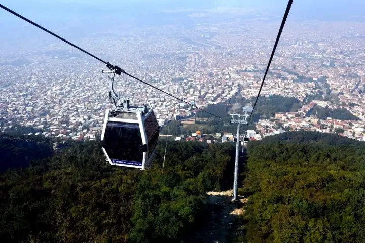 A gondola of the Bursa teleferik hangs above wooded slopes on its way down, with the rooftops of the city stretching to the horizon under a hazy sky.