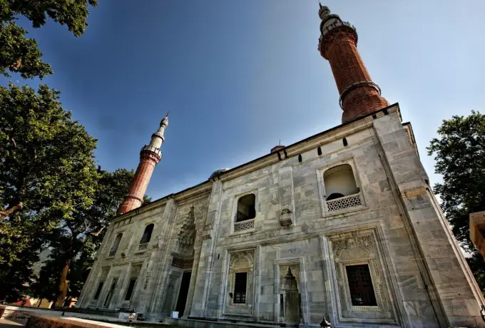 The pale stone facade of the Grand Mosque in Bursa rises against a deep blue sky, its two minarets flanking rows of arched windows and a carved doorway.