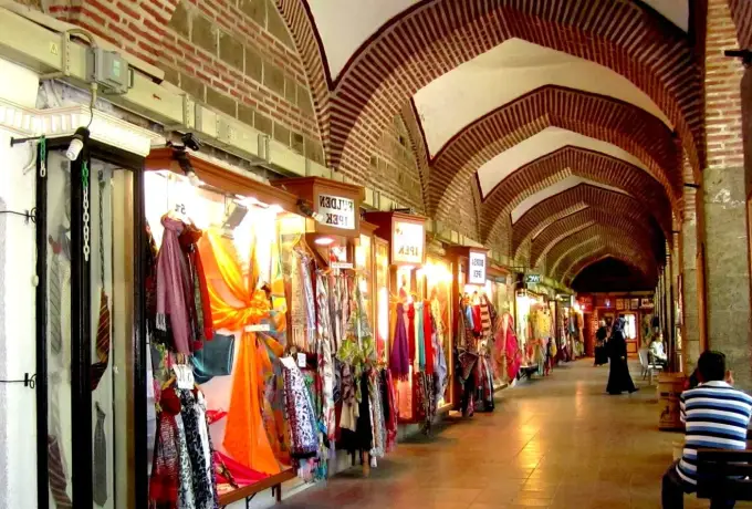 Shops selling scarves and textiles line the brick-vaulted arcade of the historic covered bazaar in Bursa, warm light falling along the polished stone floor.