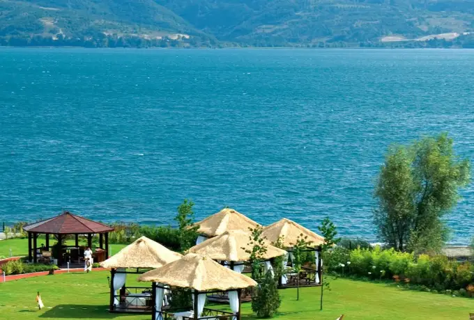 Thatched parasols and a wooden gazebo stand on a lakeside lawn at Sapanca facing the wide blue water and far hills.