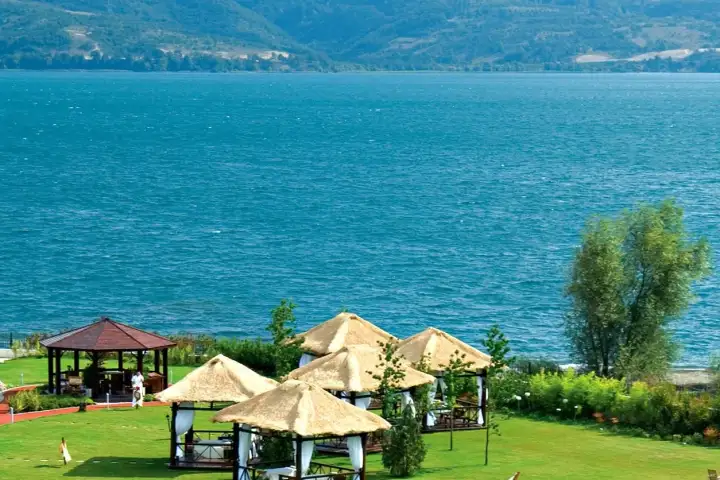 Thatched parasols and a wooden gazebo stand on a lakeside lawn at Sapanca facing the wide blue water and far hills.