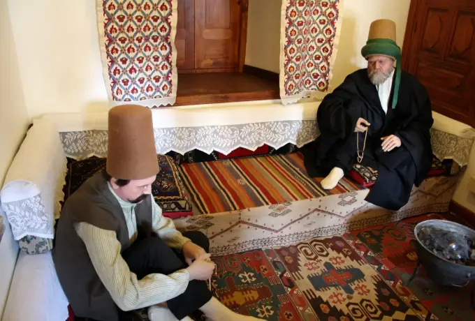Two figures in tall felt hats sit on kilim rugs in a reconstructed dervish lodge room at the Mevlana museum in Konya.