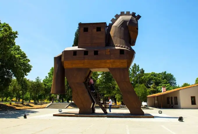 A modern angular Trojan horse sculpture in dark timber stands on a paved square beside the Troy site buildings.