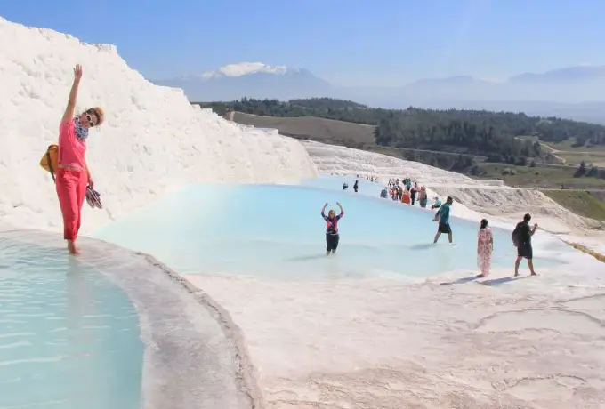 Visitors wade in the shallow blue pools of the white travertine terraces at Pamukkale under a wide summer sky.