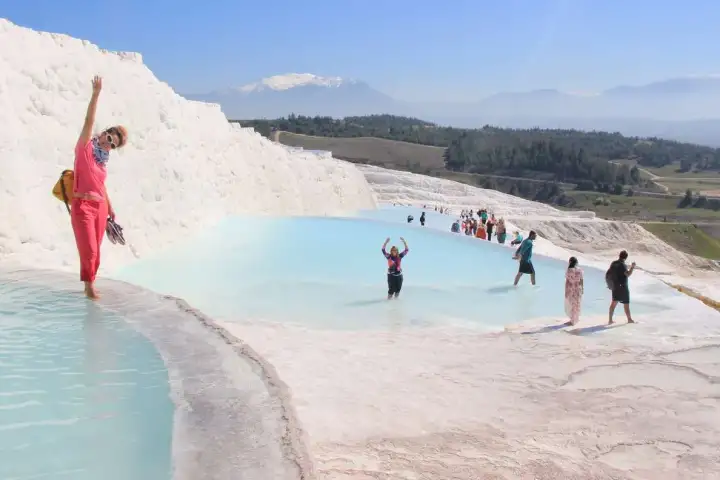 Visitors wade in the shallow blue pools of the white travertine terraces at Pamukkale under a wide summer sky.